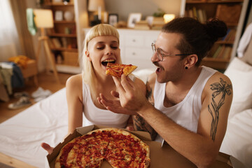 Tattooed man and his girlfriend eating pizza in bed