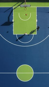 Vertical Screen: Vertical Screen: Aerial footage shows players on a blue and green basketball court from above, engaging in dynamic game. The teamwork creates vibrant scene of competition and strategy