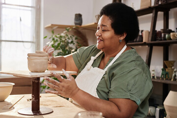 Side view portrait of senior African American woman enjoying pottery class in cozy art studio and...