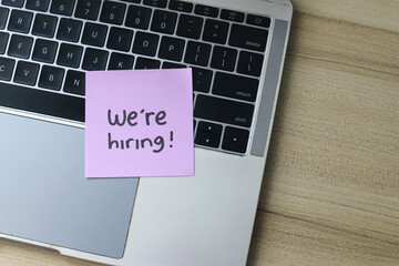 Flat Lay Of Words We're Hiring Written On Sticky Note On Keyboard Over Wooden Background.	