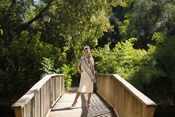 Young, beautiful, blonde woman in white suit, diamond crown and beauty pageant winner's sash, posing in the middle of a wooden bridge. Concept beauty, contests, pageant, fashion.