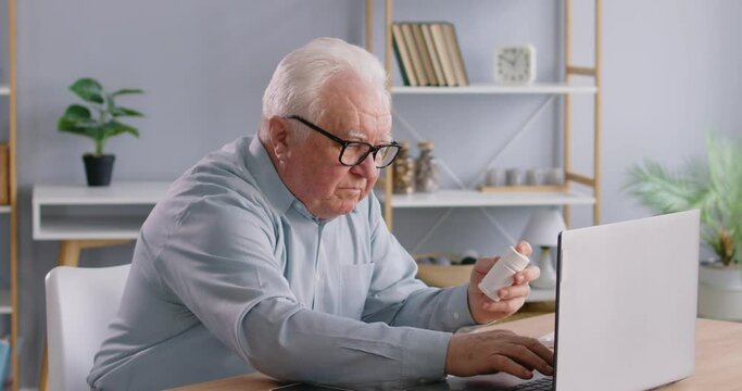 Focused senior man searching for medication instructions online for safe consumption. Pensioner holds bottle of pills and uses laptop to search for instructions for medication on Internet. 4K video.