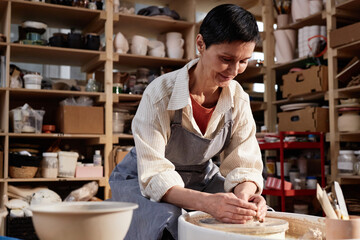 Portrait of smiling mature woman creating handmade ceramics on pottery wheel and enjoying art session in sunlight copy space