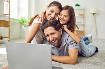 Portrait of happy cheerful young family with child girl lying on the floor at home using laptop....