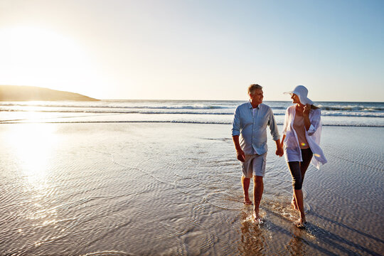 Mature couple, holding hands and walking with beach sunset for love, embrace or bonding together in nature. Man, woman or lovers enjoying stroll on water for outdoor holiday or summer by ocean coast