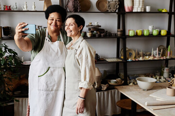 Portrait of two mature women taking selfie photo together in pottery studio, copy space
