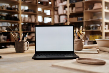 Background image of open laptop with white screen mockup on wooden table in pottery workshop, copy...