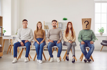 Naklejka premium Confident smiling people job candidates and seekers wearing casual clothes sitting in a row with unknown faceless man in paper bag on head with question mark. Employment and recruitment concept