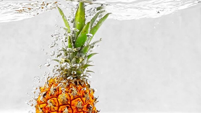 Ripe pineapple being dropped into a crystal clear water filmed in slow motion on white background. Liquid creates splashes when exotic fruit submerges showcasing freshness and juiciness. Tropical food