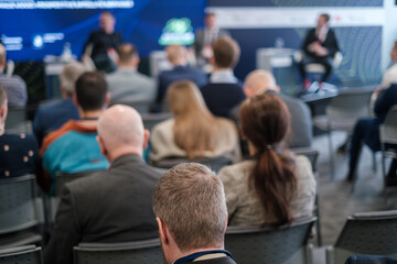 Audience at a business conference attentively listening to speakers on stage. Focus on the back of attendees' heads.
