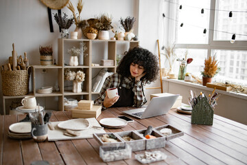 Female entrepreneur managing online order of mug on website. Young ceramic shop owner using laptop...