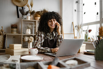 Young curly woman potter taking order for purchase of tableware using phone and laptop. Happy...