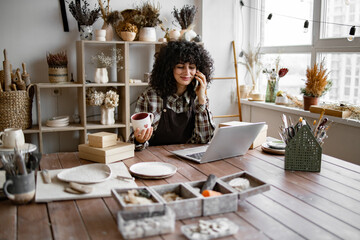 Happy female with new purchase in ceramics shop sitting at table against background of shelves with...