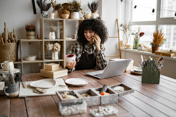 Happy female with new purchase in ceramics shop sitting at table against background of shelves with dishes. Young curly woman potter taking order for purchase of tableware using phone and laptop - Powered by Adobe