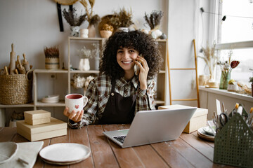 Caucasian female potter entrepreneur talking to buyer of handmade mug sitting at table and taking order using laptop. Successful business lady owner of shop made of clay talking to customer.