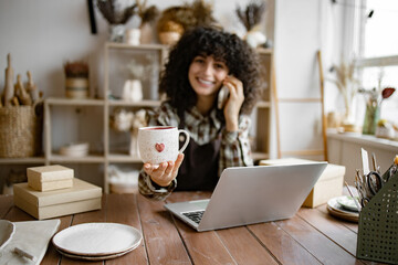 Successful business lady owner of shop made of clay talking to customer. Caucasian female potter entrepreneur talking to buyer of handmade mug sitting at table and taking order using laptop. - Powered by Adobe