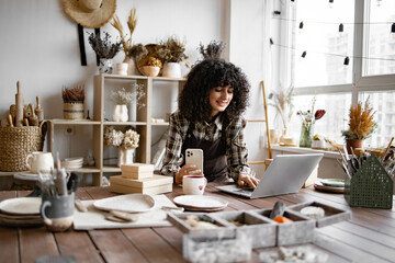 Caucasian female updates web page and social media portfolio. Curly young woman potter takes photos of handmade mugs from clay for website of her own pottery shop.