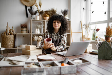 Owner of pottery workshop updates database of items that sold on website, sitting at table against...