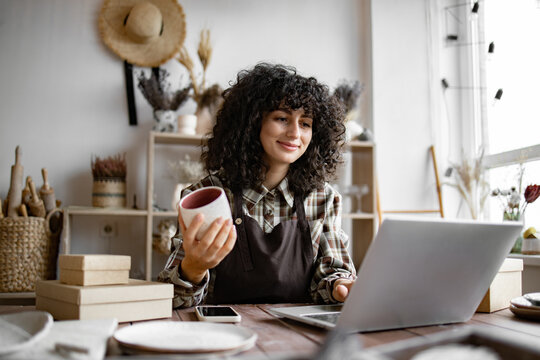 Owner of pottery workshop updates database of items that sold on website, sitting at table against background of shelves with products. Caucasian businesswoman engaged in filling social networks. - Powered by Adobe