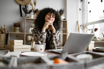 Relaxing woman in apron and checkered shirt enjoys break while sitting at table in pottery shop. Happy female saleswoman waiting for customers on background of shelves with pottery products.