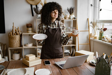 Attractive curly woman wearing brown apron receiving goods at modern decor store. Professional seller checking for no damages on cup and plate that holding in hands.