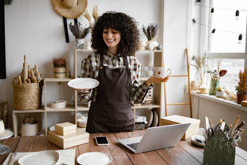 Attractive curly woman wearing brown apron receiving goods at modern decor store. Professional...