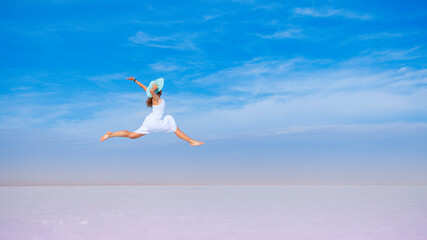 Female in white dress and blue hat jumping on blue sky- freedom, active, healthy, happiness concept