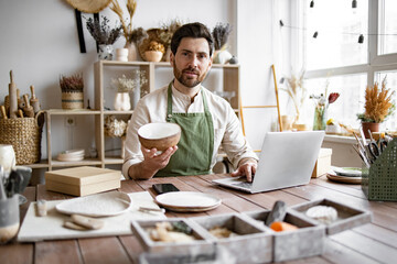 Bearded adult businessman engaged in retail trade of handmade tableware. Young Caucasian man potter happy with online sale of bowl on Internet against background of shelves with ceramics.