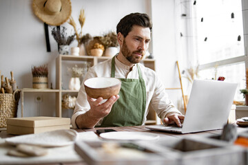 Young Caucasian man potter happy with online sale of bowl on Internet against background of shelves with ceramics. Bearded adult businessman engaged in retail trade of handmade tableware.