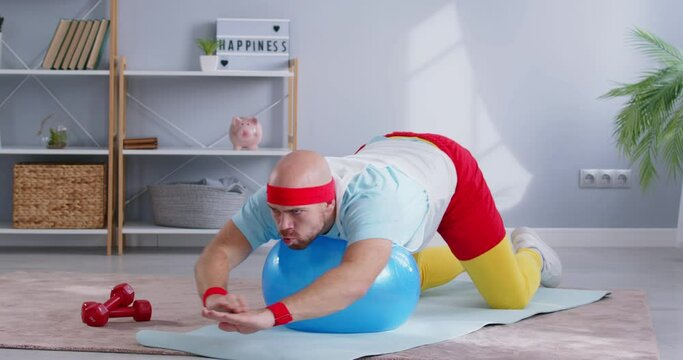 Funny young sporty bearded man wearing bright sportswear doing stretching exercises with fit ball lying on yoga mat at home. Sport, fitness, health and home workout concept. 4k video.