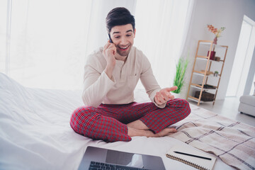 Photo of happy nice young man working from home sitting in bedroom comfy bed in white room indoors