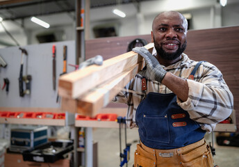 Male carpenter carrying wood planks on his shoulder. Multiracial woodworker wearing safety apron and gloves working in furniture builder and renovation in carpenter's shop. Joiner, repairman at work.