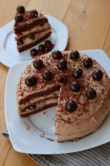 Top view of sliced  chocolate cake with cocoa cream and sour cherries in syrup decoration on a white plate on wooden table