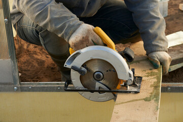 Greenhouse farming. A man cuts a board with a circular saw to strengthen a bed in a greenhouse.