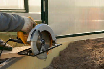 Sawing a board on the threshold of the greenhouse. A man's hand with a circular saw at work.
