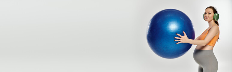 A sporty pregnant woman balances while holding a large blue fitness ball on a grey background.