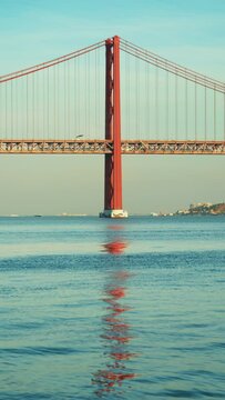 Vertical Screen: A stunning vertical video of Ponte 25 de Abril in Lisbon, Portugal, captures bridge reflected in tranquil waters under clear blue sky, showcasing this remarkable architectural wonder