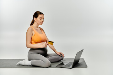 A pregnant woman in activewear sits on a yoga mat while working on a laptop.