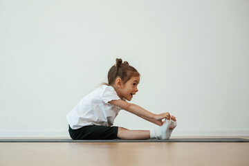Naklejka premium Side view, stretching. Cute little girl is sitting on the yoga mat and doing exercises