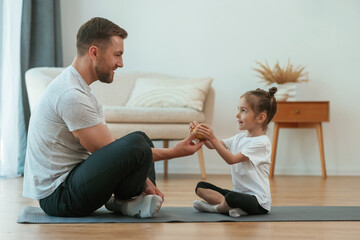 Giving an apple. Father with little daughter are doing yoga at home