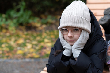 A small beautiful sad girl in glasses and a hat sits on a bench in the park in late autumn