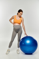 A young, sporty pregnant woman in active wear exercises next to a fitness ball on a grey background.