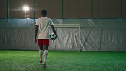 Young African American soccer player walking on artificial field indoors, holding football ball in hand, back view. Professional sport and sportive hobby, black guy training in soccer club, forward