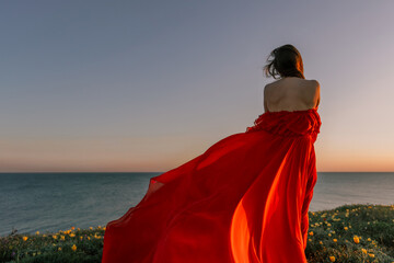 woman red dress standing grassy hillside. The sun is setting in the background, casting a warm glow...
