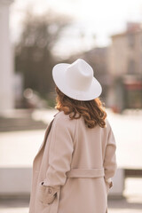 Back view of happy woman wearing hat and coat walking down street on sunny spring day. People,...