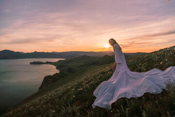 A woman in a long white dress is standing on a hill overlooking a body of water