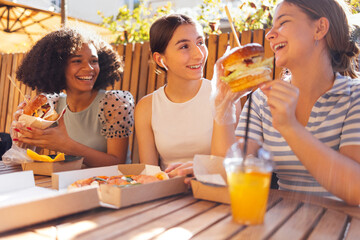 Cute smiling teenage girls are sitting in open air cafe and eating fast food