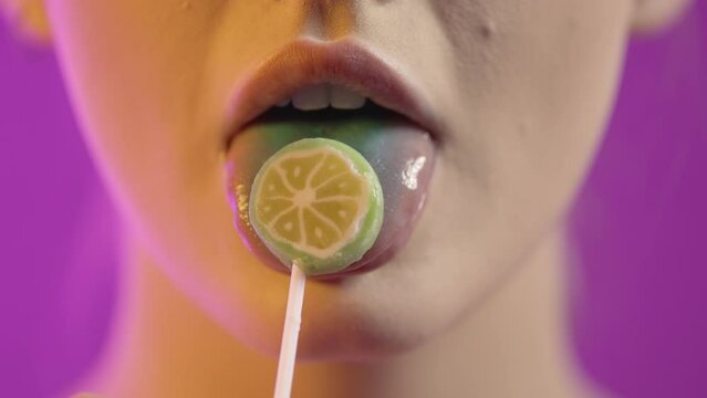 Makro ujęcie kobiecych ust na fioletowym tle. Kobieta oblizuje lizaka w kształcie plasterka pomarańczy, odsłaniając niebieski język. Gesty zmysłowe, ponętne, zakończone lekkim uśmiechem