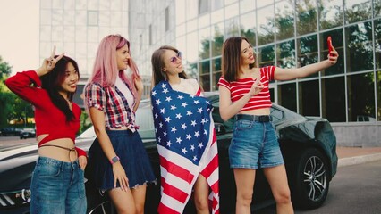 Group of beautiful multiethnic girls holding the usa flag in their hands against the background of a classic American car and take selfie and celebrate independence day. American patriot women.