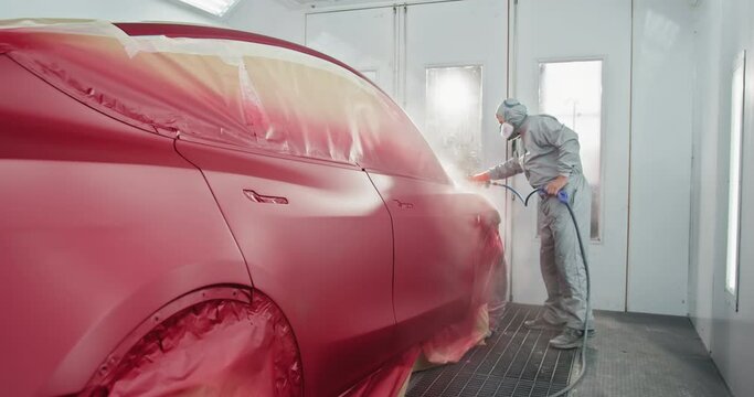A skilled worker in protective gear spray painting a red car inside a professional automotive paint chamber at a car service station. Car Painter Applying Paint to a Vehicle.